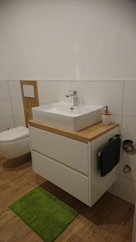 Einzelwaschtisch in weiß, aufgesetzt auf einer hellen Holzplatte mit Handtuchhalter an der Seite. 